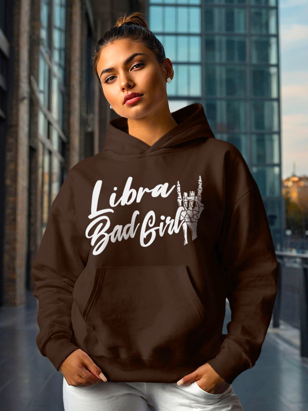 Woman wearing a dark cocoa color custom graphic hoodie with bold white lettering. The design reads “Libra Bad Girl” in script style font, with an illustration of a skeleton hand making a rock gesture next to the text. 