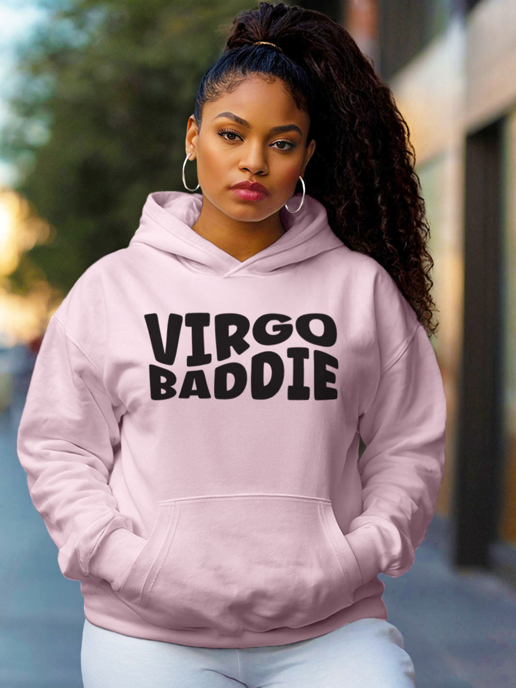 Woman wearing a light pink color custom graphic hoodie with bold black text design artwork that reads “VIRGO BADDIE” in large, playful bubble style block letters. 