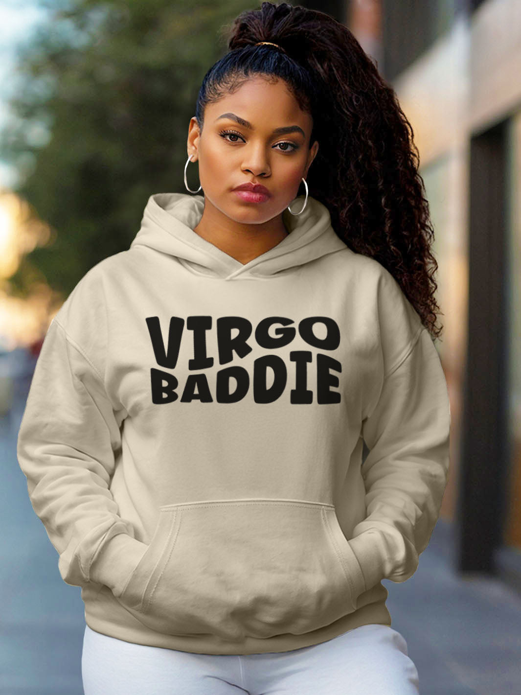 Woman wearing a sand color custom graphic hoodie with bold black text design artwork that reads “VIRGO BADDIE” in large, playful bubble style block letters. 